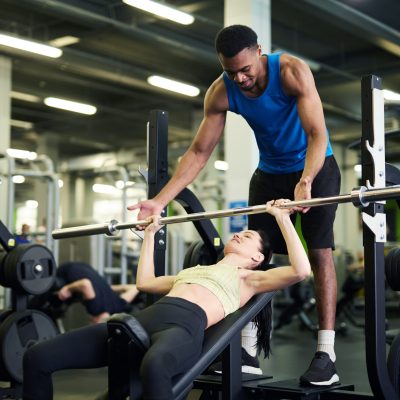 Young African-american trainer helping active girl during exercise with bar on sports equipment