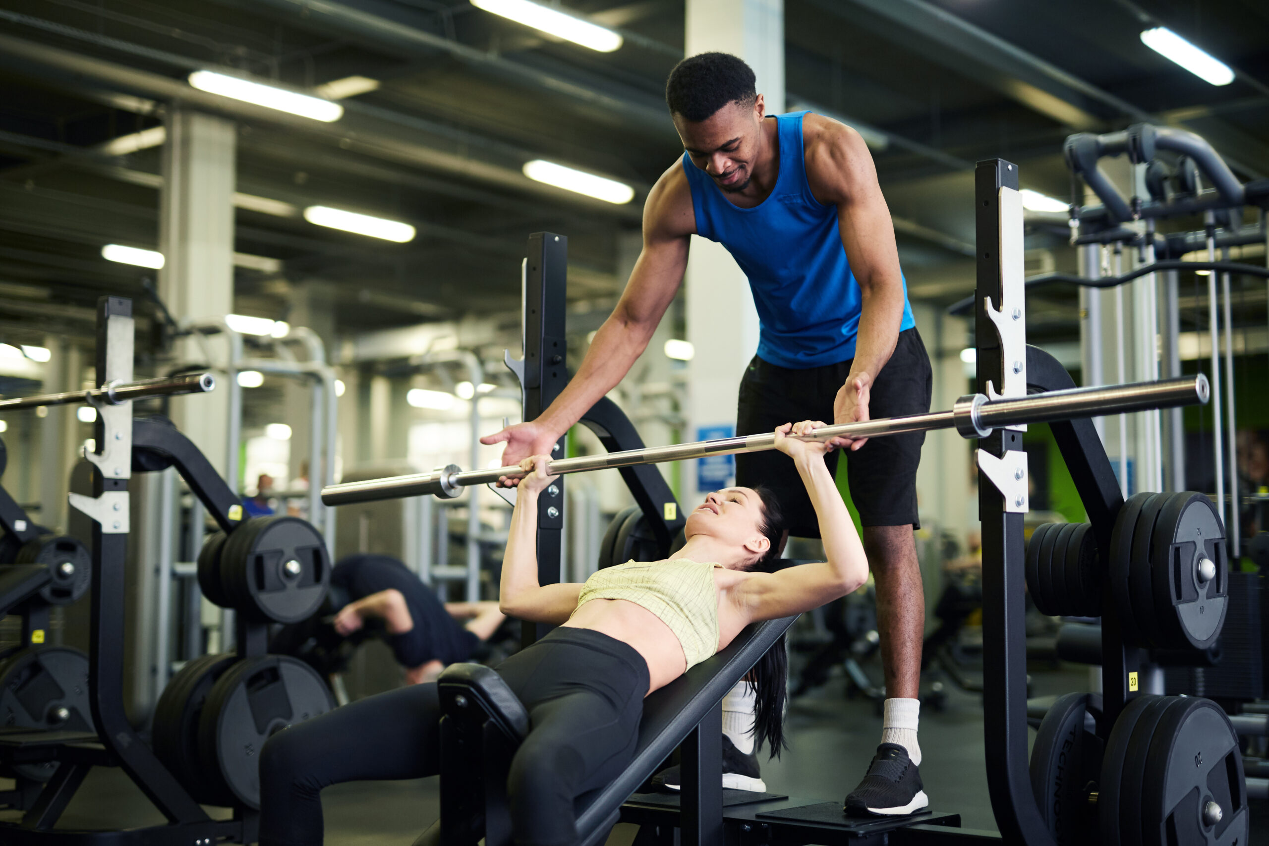 Young African-american trainer helping active girl during exercise with bar on sports equipment